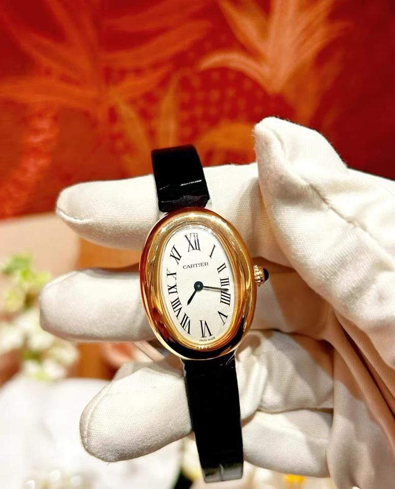 Montre ovale de teinte dorée, cadran blanc et bracelet noir, tenue avec un gant